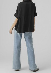 Donna che indossa una camicia nera a maniche corte larga, jeans azzurri a gamba larga e scarpe nere, in piedi contro uno sfondo grigio semplice.