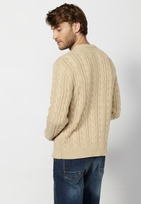 Maglione beige lavorato a maglia con motivo a treccia, scollatura rotonda e polsini a coste, abbinato a jeans blu scuro.
