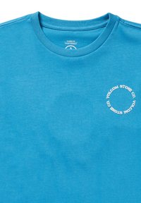 T-shirt en coton bleu avec un col rond classique et un petit logo circulaire blanc sur la poitrine gauche. Texture lisse, manches courtes.