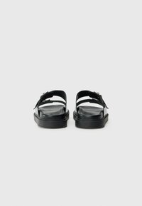 Sandales noires avec une semelle plate, des doubles sangles ajustables et une finition texturée sur le matériau, vues de l'arrière.