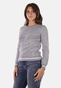 Maglione grigio con texture e motivo a onde, con polsini e orlo a coste, che presenta un piccolo emblema vicino al fondo. Abbinato a jeans blu.