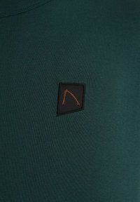 Mörkgrön kortärmad skjorta med en kvadratisk svart lappar och en orange grafisk kontur. Tyget verkar slätt och texturerat.