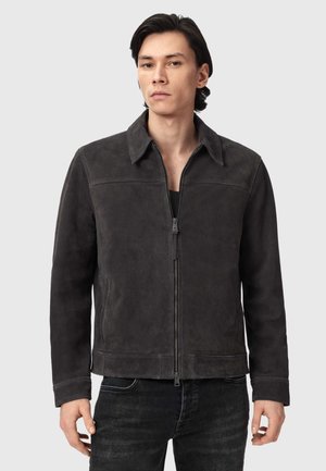 Man die een donkergrijze suède jas met ritssluiting en kraag draagt, en zwarte broek, staand tegen een vlak lichte achtergrond.