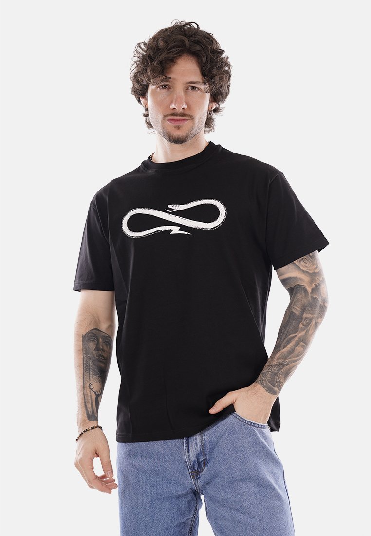 Uomo con capelli ricci che indossa una maglietta nera con simbolo di infinito a forma di serpente bianco, jeans blu e tatuaggi sulle braccia, in piedi con una mano in tasca.