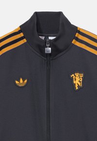 Svart zip-jacka i slät tyg. Har orange och svarta ränder på axlarna och broderade logotyper på bröstet. Ribbad krage.