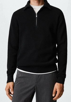 Homme portant un pull noir à manches longues avec une fermeture éclair à mi-hauteur, par-dessus une chemise blanche et un pantalon gris, debout les bras détendus le long du corps.