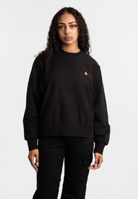 Zwarte sweater met geribde mouwen en zoom. Heeft een rood en geel logo op de linkerkant van de borst. Materiaal lijkt zacht en gestructureerd.