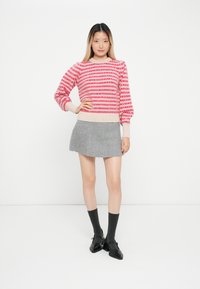Sweter dzianinowy w czerwone i różowe paski, z prążkowanymi mankietami, zestawiony z szarym minisukienką. Modelka nosi szare skarpetki i czarne buty, stojąc na białym tle.