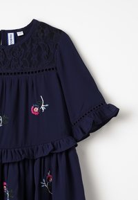 Granatowa sukienka z haftowanymi kwiatowymi detalami w różowym i zielonym kolorze. Zawiera koronkowe akcenty na dekolcie i falbaniaste rękawy. Wykonana z lekkiego materiału.