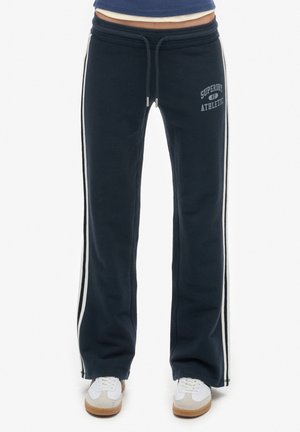 Pantalon de sport bleu marine avec bandes blanches latérales, taille à cordon, logo « Superdry Athletic » sur la cuisse, porté avec des baskets blanches.