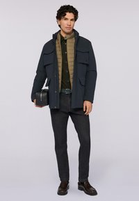 Uomo che indossa una giacca blu navy a strati, un gilet imbottito verde oliva, jeans scuri e mocassini marroni, mentre tiene una borsa rettangolare nera su uno sfondo semplice.