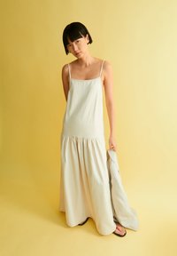 Helles Beige, ärmelloses Maxikleid mit eng anliegendem Oberteil und gerafftem Rock, aus weichem Stoff gefertigt. Das Model hält eine leichte Jacke.