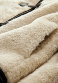 Beige pūkains audums ar mīkstu, reljefu virsmu. Iekļauj melnu rāvējslēdzēju un ādas akcentus. Tuvplāns, kas izceļ materiāla detaļas.