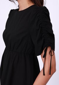 Vestido negro con un corpiño ajustado y mangas abullonadas, que presenta detalles de elasticidad recogida y lazos ajustables en los puños. Textura de tela suave.