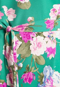 Vestido floral com um fundo verde vibrante e flores em tons de rosa, branco e azul. Inclui um detalhe de cintura amarrada com uma textura subtil.