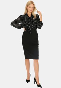 Czarna bluzka w kropki z wiązanym dekoltem, w zestawieniu z dopasowaną czarną spódnicą. Strój ma długie rękawy i czarne buty na wysokim obcasie.