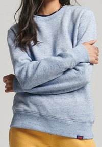 Persona che indossa un maglione azzurro chiaro a pois e pantaloni giallo senape, con le braccia incrociate, ritagliata al collo e ai fianchi.
