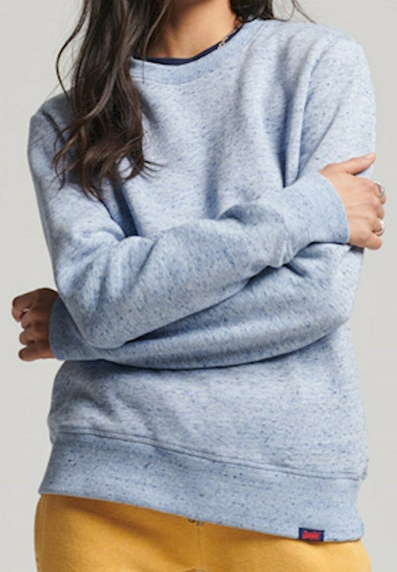 Persona che indossa un maglione azzurro chiaro a pois e pantaloni giallo senape, con le braccia incrociate, ritagliata al collo e ai fianchi.