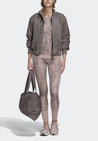 Žena v legínách a topu s hadím potiskem, v šedé bomber bundě, drží velkou šedou tašku, stojí na jednobarevném pozadí.