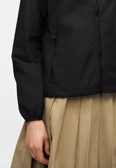 Bras gauche et torse vêtus d'une veste noire à fermeture éclair avec des poignets élastiques, portée sur une jupe plissée beige, sur un fond uni.