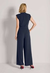 Vrouw met kort blond haar staat met haar rug naar de camera, gekleed in een mouwloos marineblauw jumpsuit en rode hoge hakken, tegen een effen grijze achtergrond.