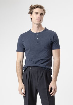 Joven con cabello castaño claro lleva una camiseta henley azul marino de manga corta y pantalones oscuros plisados, de pie frente a un fondo liso.