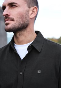 Svart knappad skjorta i mjukt tyg, med krage och en liten logotyp på bröstet. Underneath finns en vit T-shirt.