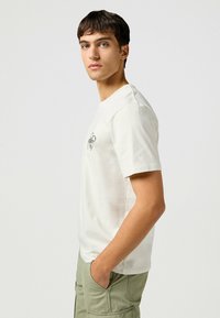 Short-sleeve, cream cotton t-shirt with a round neckline and black embroidered graphic on the chest, paired with green cargo pants.