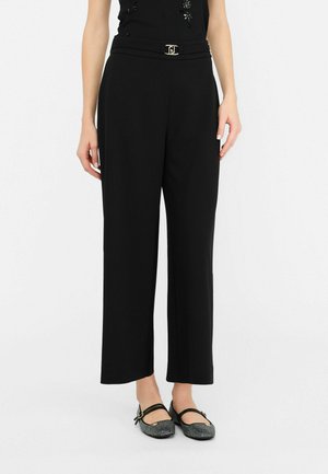 Schwarze weit geschnittene Hose mit glatter Textur, die eine taillierte Taille und ein metallisches Detail aufweist. Kombiniert mit flachen, gemusterten Schuhen.