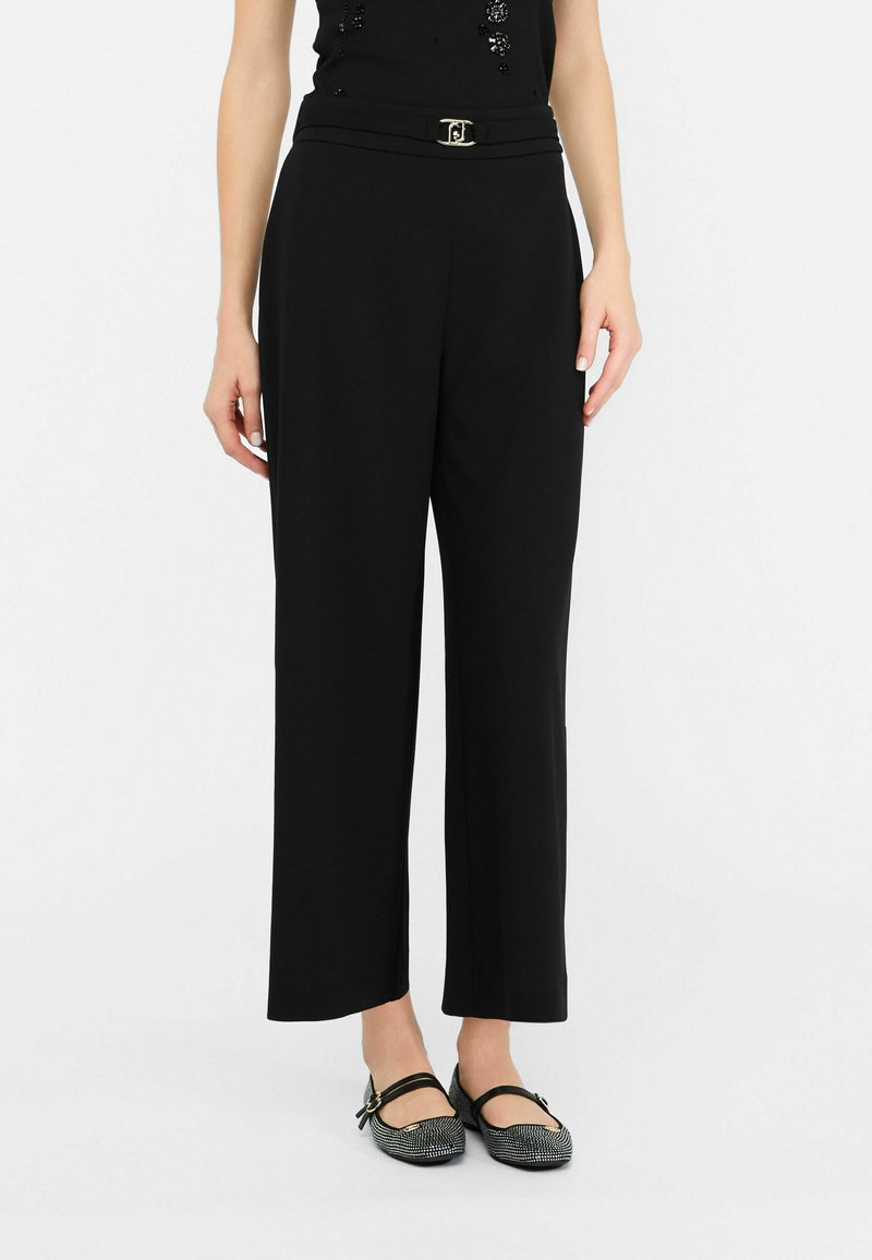 Zwarte wijde broek met een gladde textuur, voorzien van een ceintuur en een metallic accent. Gecombineerd met platte, gemusterde schoenen.