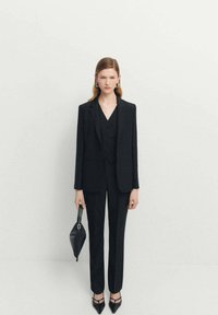 Čierny oblek na mieru so sakom na jeden gombík, zodpovedajúcim vestom a úzkymi nohavicami. Doplní ho malá čierna kabelka a špicaté lodičky.