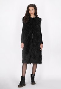 Schwarze Fleece-Weste aus Kunstfell mit einem langen, fließenden Design und einem Reißverschluss vorne. Trägt sie über einem schwarzen Langarmtop und kombiniert mit schwarzen Schnürstiefeln.