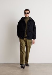 Sort hættetrøje med store lommer, kombineret med en tætsiddende olivengrøn turtleneck og khaki bukser, båret med mørke solbriller og sorte sko.