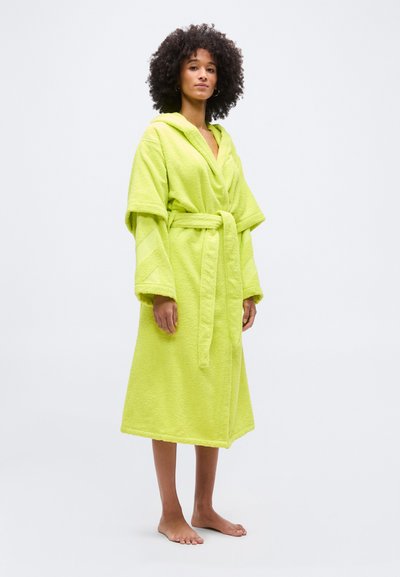 Femme aux cheveux bouclés portant un peignoir à capuche vert citron vif, noué à la taille, debout pieds nus sur un fond clair uni.