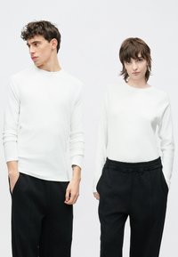 Valged pika varrukaga pealsed, mida kantakse mustade pükstega. Meeste pealsel on ümara kaelusega kampsun; naiste pealsel on ümar kaelus ja lõdvestunud lõige.