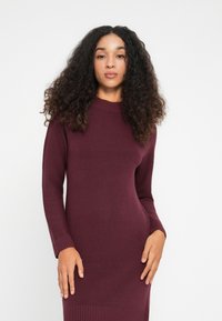 Donna con capelli ricci che indossa un vestito a maniche lunghe in maglia a coste color borgogna, in piedi davanti a uno sfondo bianco uniforme.