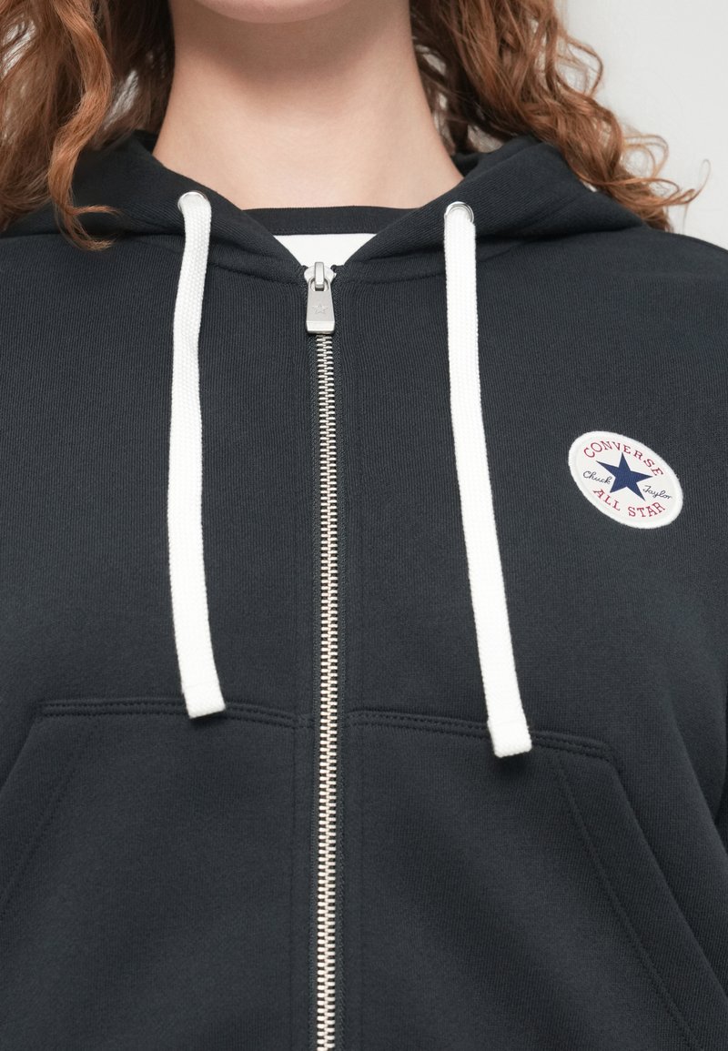 Sort lynlås-hættetrøje lavet af bomuld. Har hvide snore og et rundt logopatch med tekst og en stjerne. Glat tekstur.