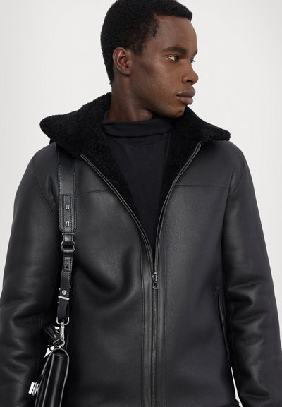 Veste en cuir noir avec un col en shearling, fermeture éclair et poches latérales ; associée à un col roulé noir et portant un sac en cuir.