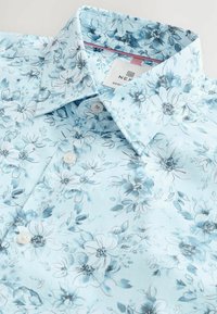Camicetta floreale azzurro chiaro con motivi di fiori bianchi e blu navy, design con colletto e chiusura a bottoni. Tessuto liscio con una finitura leggermente lucida.