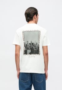 Camiseta de algodón blanca con un gráfico de paisaje urbano en blanco y negro en la parte posterior, enmarcado por texto en diferentes tipos de letra. Mangas cortas y corte relajado.