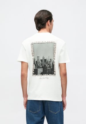 T-shirt en coton blanc avec un graphique monochrome de paysage urbain au dos, encadré par du texte en différentes polices. Manches courtes et coupe décontractée.