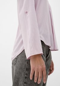 Camicia a maniche lunghe rosa chiaro con orlo arrotondato e dettaglio a bottone sulla manica, abbinata a pantaloni in denim grigi. Texture del tessuto liscia.