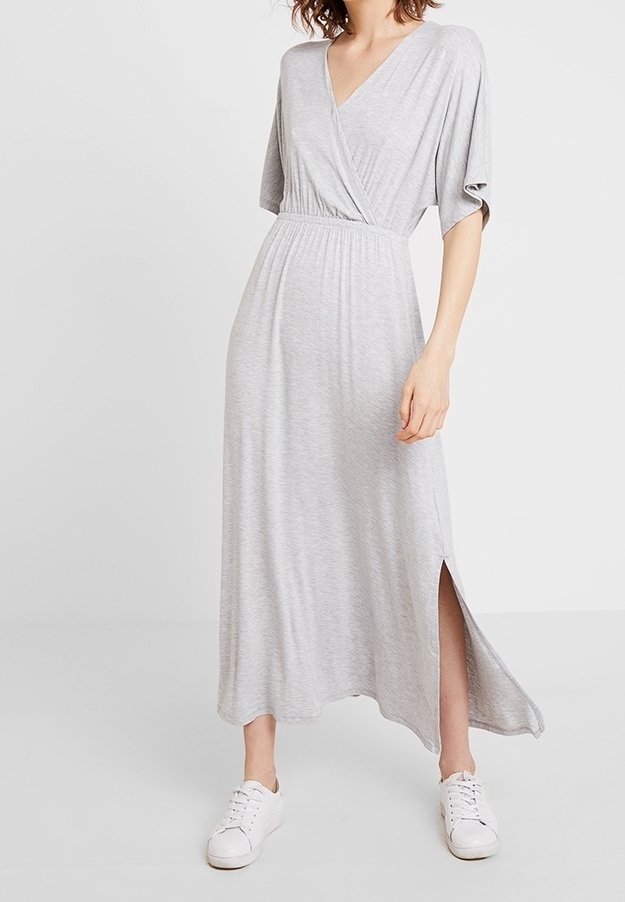 Femme portant une robe maxi gris clair style portefeuille avec manches courtes et fente latérale, associée à des baskets blanches, debout devant un fond uni.