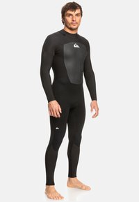 Man staat blootvoets in een zwart wetsuit met volledige bedekking en Quiksilver-logo's op de borst en bovenbeen tegen een effen lichte achtergrond.
