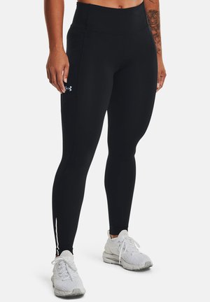 Piernas con leggings negros de cintura alta con el logo de Under Armour, combinados con zapatos deportivos blancos sobre un fondo blanco liso.