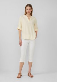 Blouse jaune clair à manches courtes avec un décolleté en V et des manches roulées, associée à un pantalon capri blanc et des sandales plates brunes. Design minimal.