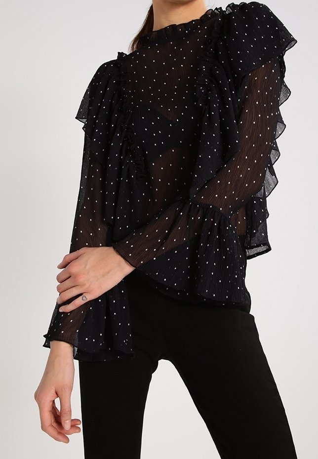 Femme portant un chemisier noir transparent à manches longues avec des pois blancs et des détails volantés, associé à un pantalon noir.