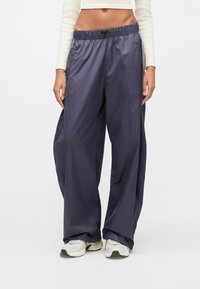 Γυναίκα που φορά χαλαρό σκούρο γκρι αθλητικό παντελόνι με ελαστική μέση και λευκά αθλητικά παπούτσια, με λευκό μακρυμάνικο κοντό τοπ που αφήνει ακάλυπτη τη μέση.