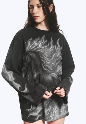 Femme portant une chemise noire à manches longues oversize avec un motif détaillé de cheval blanc et de flammes, regardant sur le côté les mains jointes devant.