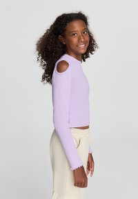 Top lungo lavanda a maniche lunghe con aperture sulle spalle, texture a coste e polsini a balze, abbinato a pantaloni bianchi con vita elasticizzata.